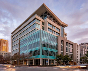 An eight story, blue and tan, life science building with a crosswalk and cars driving in front of it.