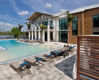 A brown and white, plaster and wood residential clubhouse on the edge of the water with a blue pool and sunbathing chairs in front of it.